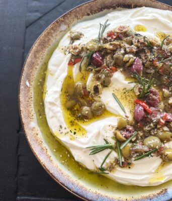 Creamy whipped feta dip topped with olive tapenade, herbs, and olive oil, served in a rustic bowl for event catering.