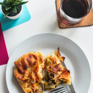 A plate with a savory pastry, partially eaten, sits next to a fork and knife. Nearby are a glass of dark red drink on a coaster and a small potted succulent on a white table with colorful mats.