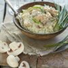 A rustic bowl of white rice topped with creamy chicken and green bell peppers, garnished with chives. Sliced white mushrooms and fresh chives are arranged beside the bowl on a wooden surface.