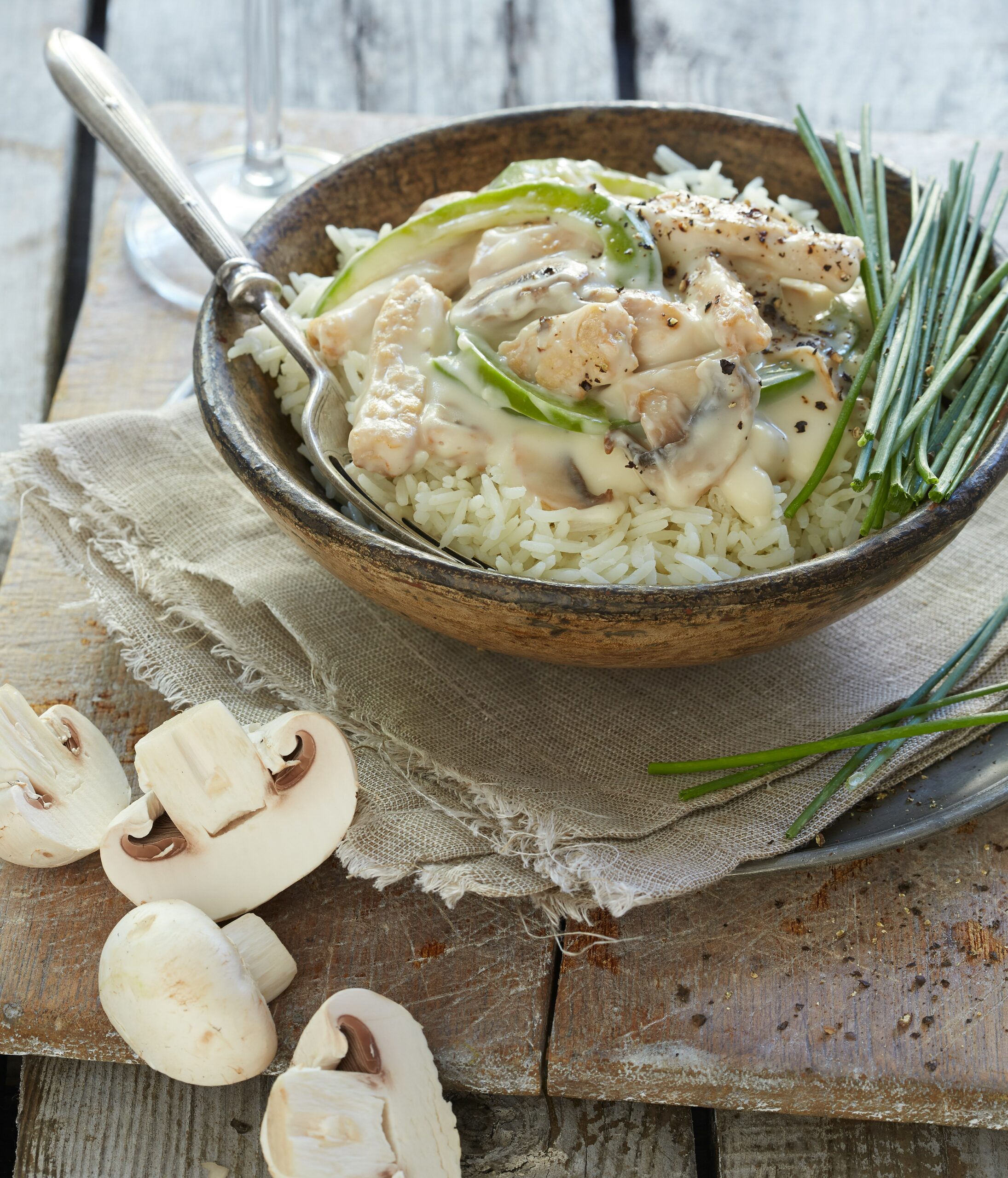 Chicken ala King A rustic bowl of white rice topped with creamy chicken and green bell peppers, garnished with chives. Sliced white mushrooms and fresh chives are arranged beside the bowl on a wooden surface.