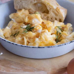 A white bowl filled with scrambled eggs garnished with fresh dill, served with a piece of bread on a wooden surface.