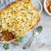 A baked shepherd’s pie topped with golden mashed potatoes sits in a casserole dish with a portion removed. Nearby, a serving is in a bowl beside a fork and spoon on a textured surface. Fresh herbs are sprinkled on top.