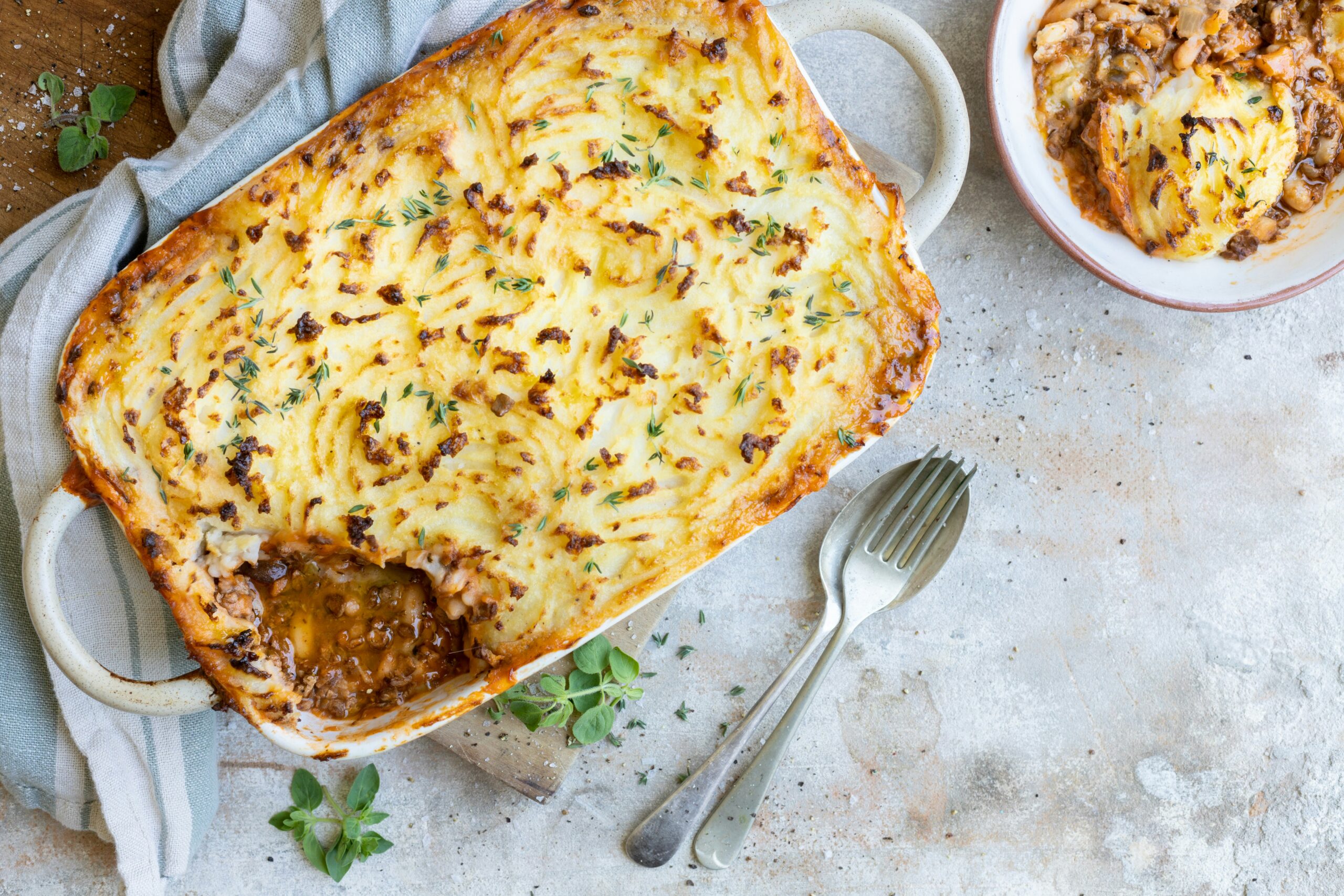 Shepards Pie A baked shepherd’s pie topped with golden mashed potatoes sits in a casserole dish with a portion removed. Nearby, a serving is in a bowl beside a fork and spoon on a textured surface. Fresh herbs are sprinkled on top.