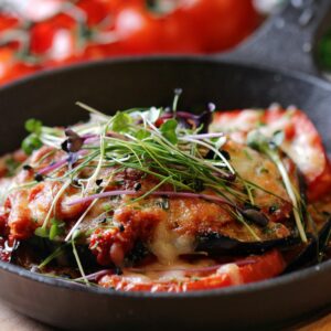 A skillet filled with baked eggplant parmesan, topped with melted cheese, tomato slices, and a garnish of fresh microgreens, with tomatoes visible in the background.